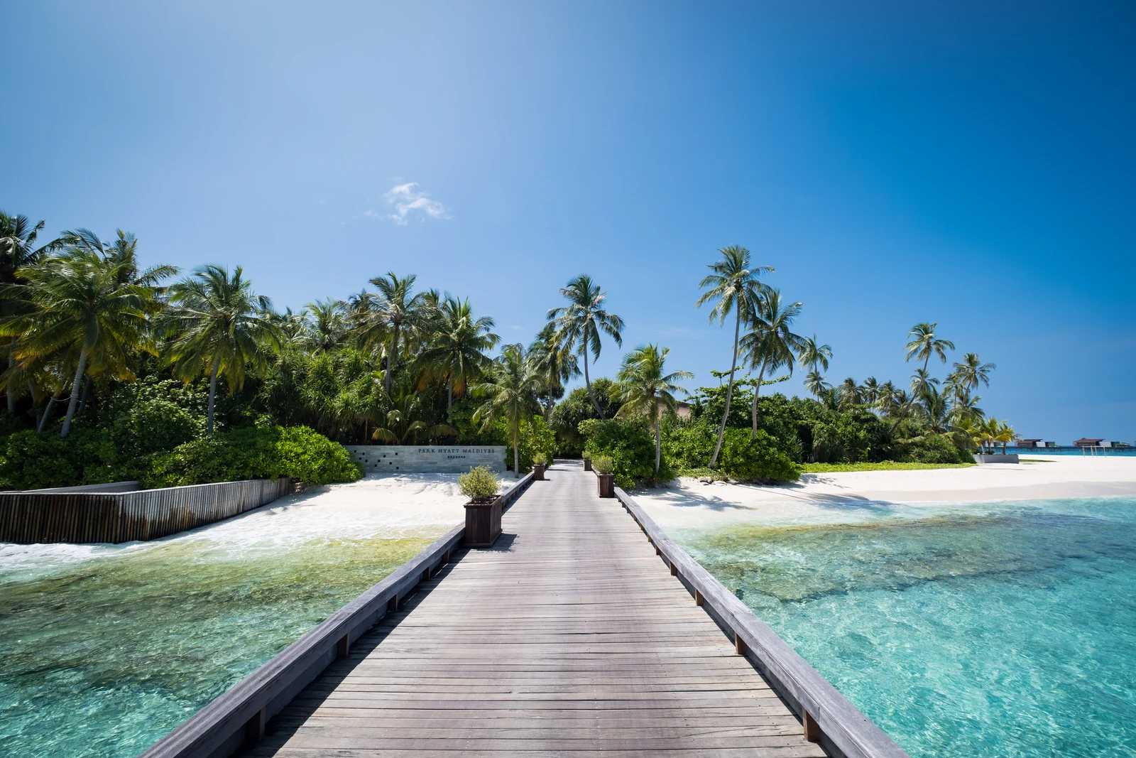 Деревянный причал ведёт к берегу Park Hyatt Maldives Hadahaa; слева видна вывеска курорта, справа — пляж и водные виллы вдали.